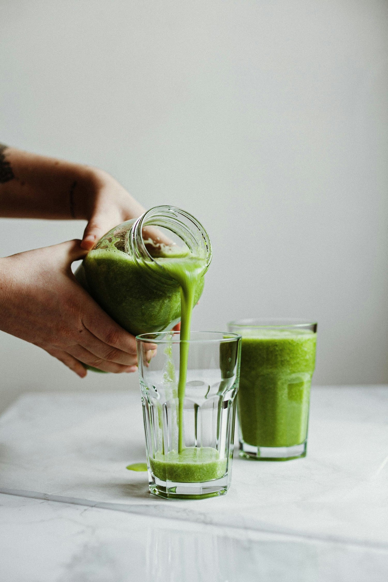 full glass of green juice set on marble countertop and woman pouring glass of green juice into another glass