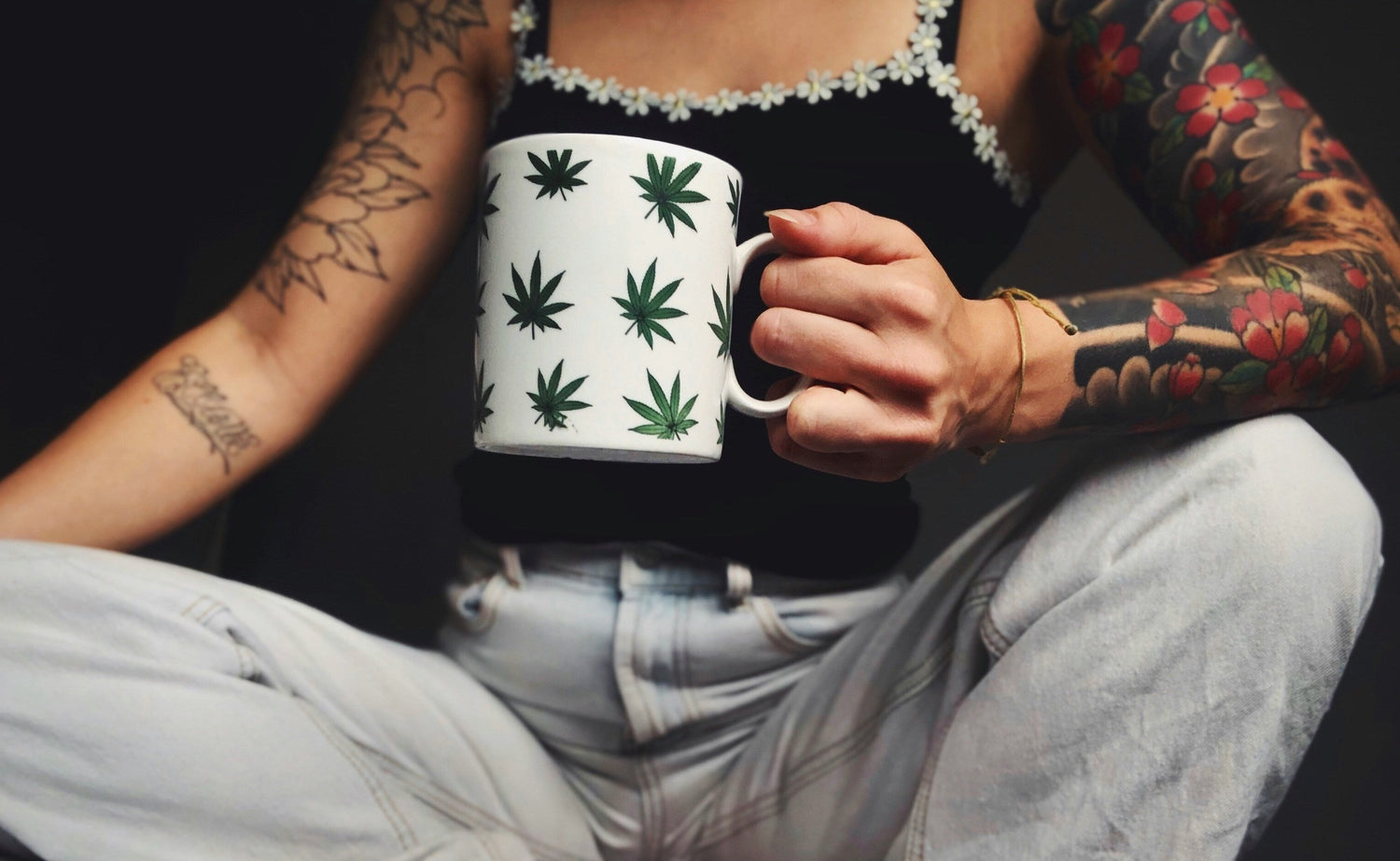 woman with arm tattoos wearing jeans and holding a white mug with green hemp plant design