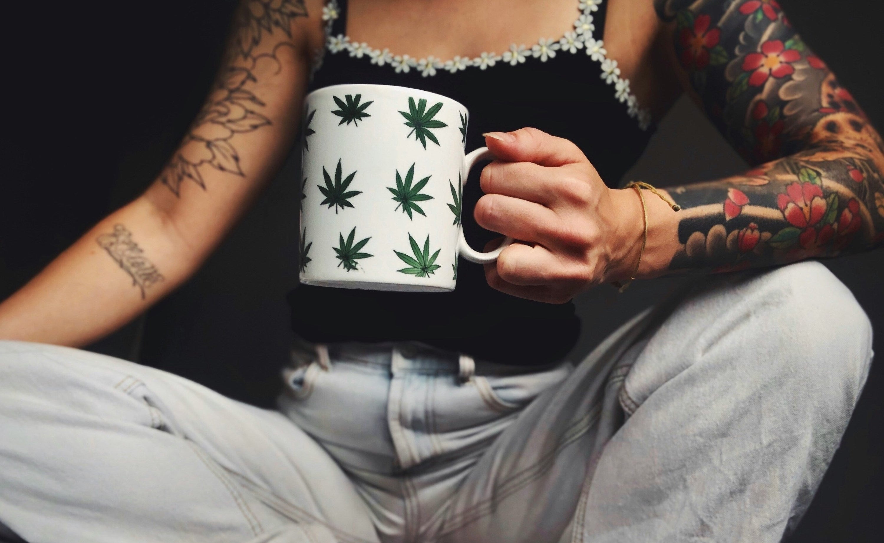 woman with arm tattoos wearing jeans and holding a white mug with green hemp plant design